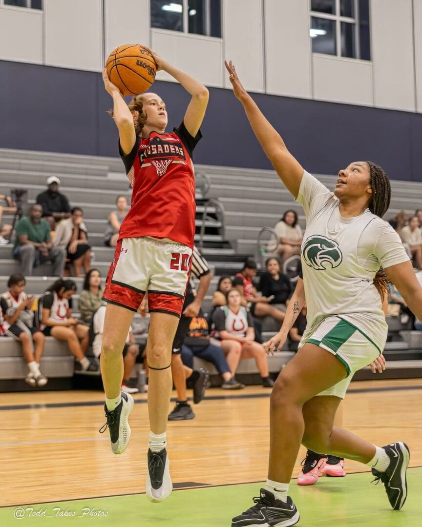 A basketball player in red jumps to shoot over a defender in white.